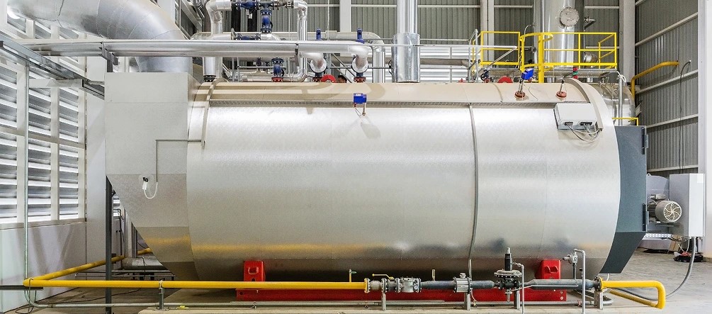 A packaged firetube industrial boiler installed in a plant boiler room, with feedwater piping and stack connections visible